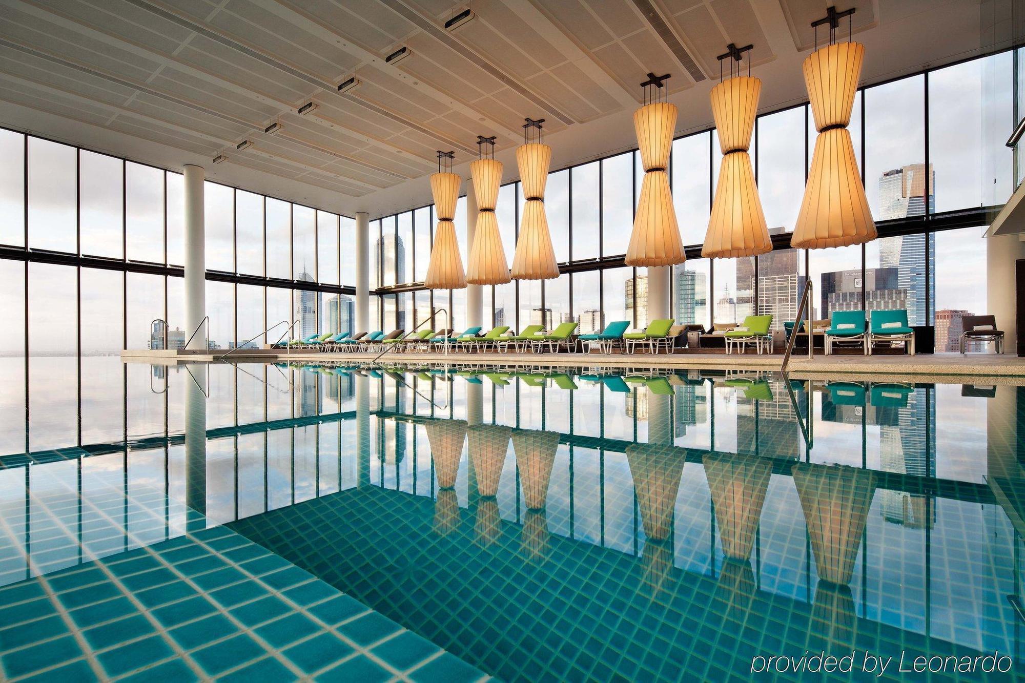 Crown Metropol Melbourne Hotel Exterior foto The photo shows a modern indoor swimming pool area. The pool features a tiled surface and reflects large, stylish light fixtures hanging from the ceiling. Surrounding the pool are several lounge chairs arranged neatly, and the walls consist of large 