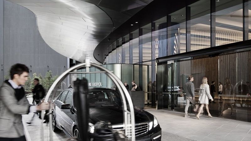 Crown Metropol Melbourne Hotel Exterior foto The image shows a modern hotel entrance with sleek architecture. In the foreground, there is a black luxury car parked near a curved entryway. A luggage cart is positioned to the side, indicating a welcoming environment for guests. People are seen wa