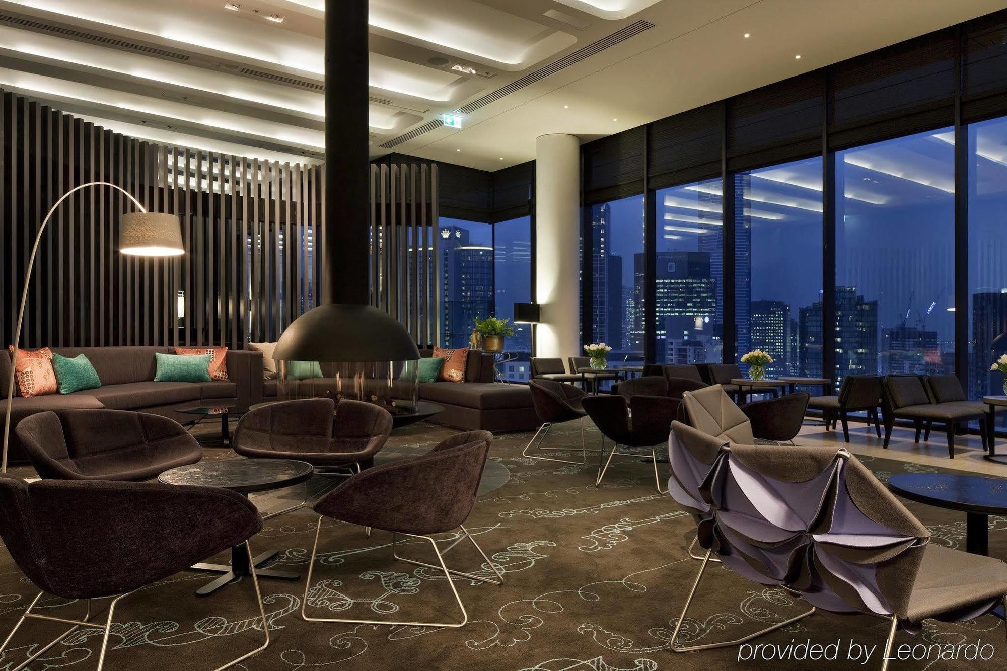 Crown Metropol Melbourne Hotel Exterior foto The image depicts a modern and stylish lounge area. The space features large windows that provide a view of a city skyline at dusk. The interior is designed with a combination of dark and light elements, including a sleek fireplace in the center. The